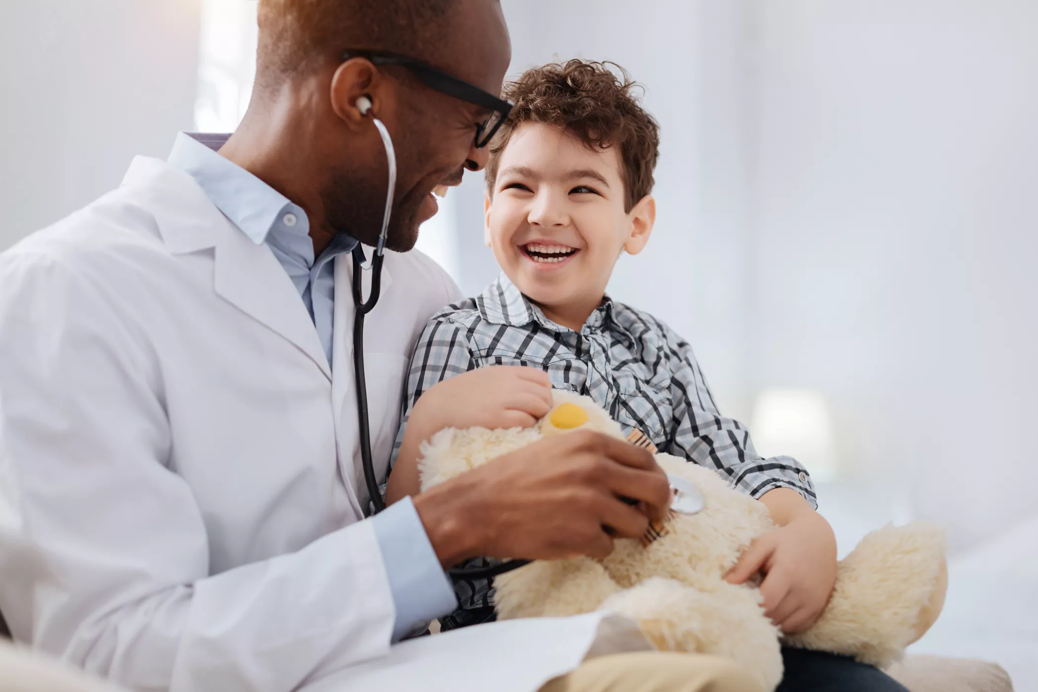 A healthcare professional in a white coat uses a stethoscope to examine a young child holding a stuffed teddy bear. The setting appears to be a bright and clean medical office or clinic. The scene conveys care, trust, and a comforting environment for pediatric healthcare.